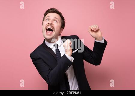 Giovane uomo d'affari caucasico con baffi che indossa la tuta danzando e cantando sulla parete rosa. Celebrazione e successo. Studio Foto Stock