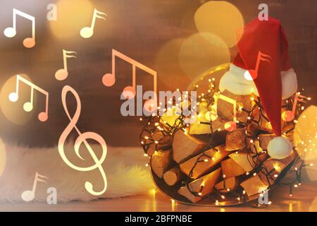 Cesto con legna da ardere, cappello Santa e garland su sfondo luci sfocate. Concetto di musica e canzoni natalizie Foto Stock