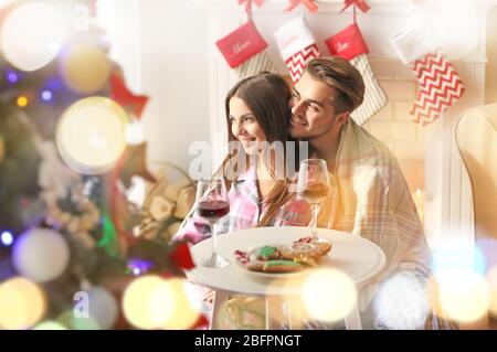 Giovane coppia seduta accanto al tavolo con vino e biscotti in camera decorata per Natale Foto Stock