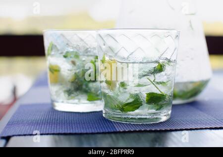 Due bicchieri pieni di acqua fresca e fresca trasparente con ghiaccio, limone e basilico foglie su tavola blu Foto Stock