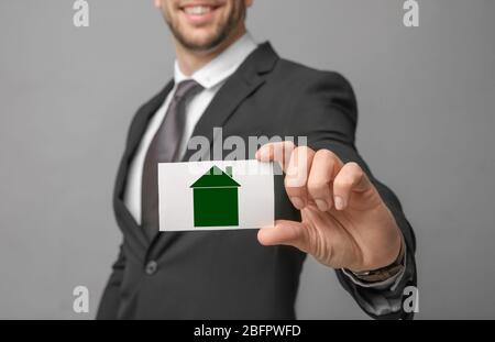 Agente immobiliare in possesso di biglietto da visita con disegno di casa su sfondo grigio Foto Stock