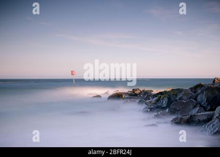 una lunga immagine di onde che si infrangono sulle rocce al tramonto con un orizzonte chiaro Foto Stock