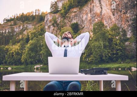 Un uomo barbuto lavora con un laptop sulla natura. Un libero professionista felice è seduto e utilizzando un'applicazione o un sito web sull'erba. Lavorare da remoto in autonomia Foto Stock