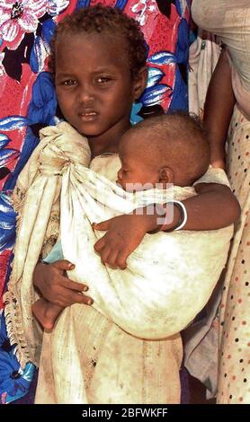 1993 - dritto, medium close-up di una giovane ragazza somala cullano la sorella in fascia che è legato sulla sua spalla. Essi sono in attesa in linea per vedere US Navy medici conducendo un medico civile Programma d'azione nella capitale Mogadiscio, Somalia. Foto Stock