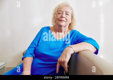 Ritratto di donna anziana in elegante abito blu scuro seduto sul divano a casa e guardando la macchina fotografica Foto Stock