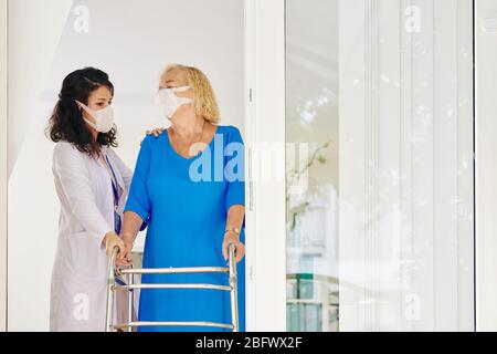 Giovane operatore sanitario che sostiene il paziente anziano che si muove con il deambulatore Foto Stock