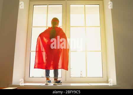 Bambina in un costume supereroe in piedi sullo sfondo di una finestra con luce del sole guardando i sogni alla finestra