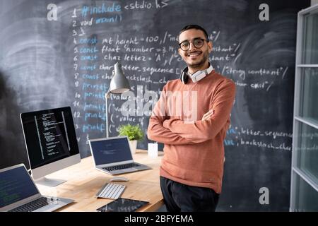 Ritratto di soddisfatto giovane programmatore medio-orientale con cuffie intorno al collo in piedi con braccia incrociate in studio di codifica Foto Stock