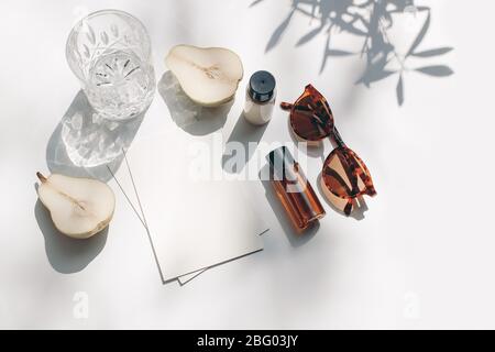 Cancelleria estiva ancora vita scena. Bicchiere d'acqua, frutta a pera tagliata, olio cosmetico, bottiglie di crema e occhiali da sole, tavolo bianco. Carta bianca c Foto Stock