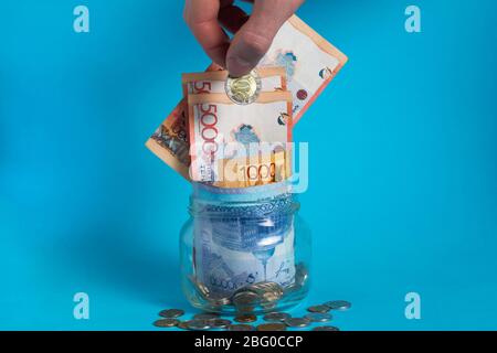La mano mette una moneta di tenge in un vaso di vetro con soldi. Kazakistan. KZ. KZT. Banconote, banconote, monete, Banca. Stipendio, credito, risparmio, paga e spe Foto Stock