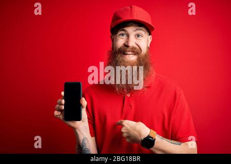 Uomo di consegna irlandese Redhead con smartphone che tiene la barba e mostra l'app sullo schermo con il viso a sorpresa rivolto a se stesso Foto Stock