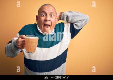 Uomo anziano bello bere vaso di birra in piedi su sfondo giallo isolato Crazy e spaventati con le mani sulla testa, paura e sorpreso di shock w Foto Stock