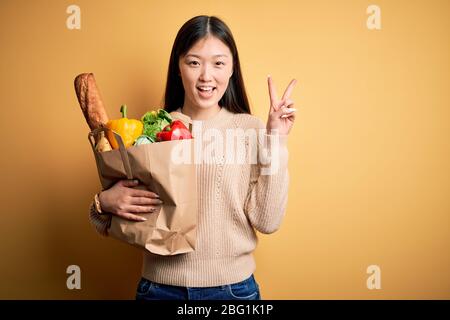 Giovane donna asiatica che tiene sacchetto di carta di generi alimentari sani freschi su sfondo isolato giallo sorridente con faccia felice che si annida alla telecamera facendo vic Foto Stock