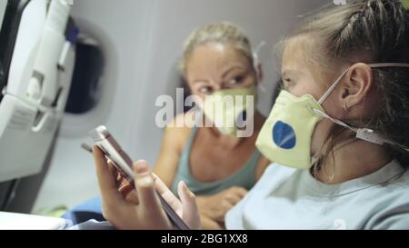 Famiglia donna e bambino viaggio turistico caucasico in aereo aereo con indossare maschera medica protettiva. Utilizzare il cellulare dello smartphone. Covid-19 Foto Stock