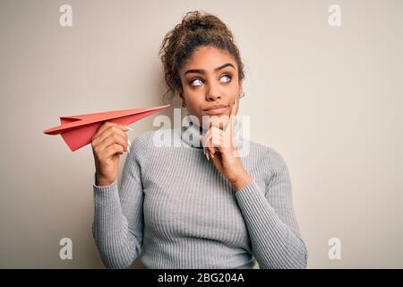 Giovane afro americano afro ragazza che tiene aereo di carta su isolato sfondo bianco serio faccia pensare alla domanda, molto confuso idea Foto Stock