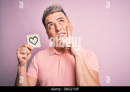 Giovane uomo romantico moderno tenendo forma del cuore disegno su sfondo rosa isolato serio faccia pensare alla domanda, molto confuso idea Foto Stock