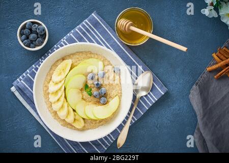 Farinata d'avena con banane, mirtilli, marmellata, miele, tovagliolo blu su fondo di pietra blu Foto Stock