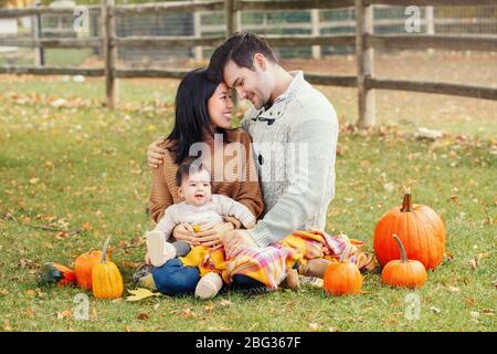 Madre cinese asiatica e padre caucasico con bambina seduta in autunno parco all'aperto. Famiglia mamma, papà e figlia in fattoria con zucche. Foto Stock