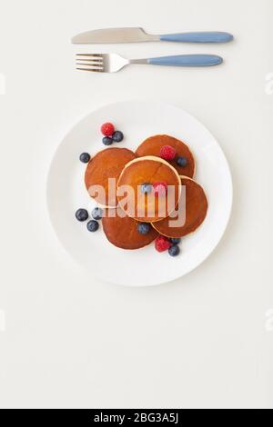 Vista dall'alto con una composizione minima di deliziosi pancake dorati decorati con bacche fresche accanto al coltello e forchetta su sfondo bianco, colazione concep Foto Stock