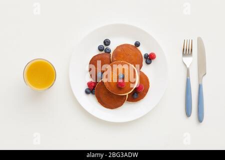 Vista dall'alto con una composizione minima di deliziosi pancake dorati con bacche fresche accanto al succo d'arancia e coltello e forchetta su sfondo bianco, colazione Foto Stock