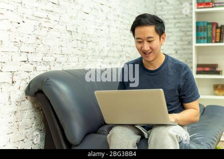 Giovane uomo asiatico sorridente in abbigliamento casual sul divano di lavoro sul computer portatile. Freelance maschio che lavora da casa su notebook. Lavoro da casa concetto Foto Stock