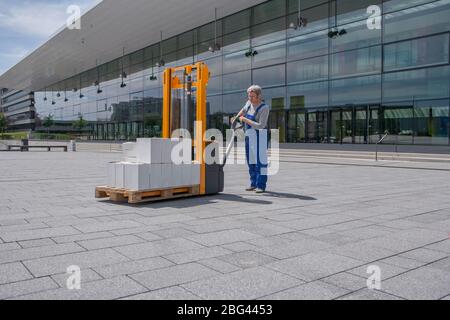 uomo in tuta generale aziona un carrello elevatore caricato con scatole Foto Stock