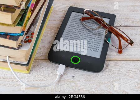 Occhiali da vista su lettore di e-book collegati via cavo a una pila di libri di carta su un tavolo di legno bianco. E-reading per piacere e istruzione. Vista dall'alto. Foto Stock