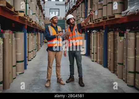 Una lunga serie di manager professionisti che lavorano insieme mentre si trovano in magazzino, tenendo il tablet digitale rivolto verso la scatola di cartone Foto Stock