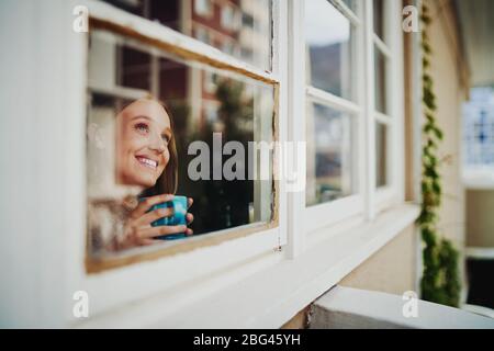 Bella giovane donna sorridente bere caffè e sognare di giorno godendo di vista esterna durante l'isolamento Foto Stock