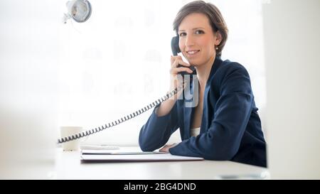 Giovane e simpatica donna d'affari che parla al telefono mentre si siede alla scrivania in ufficio girandosi per sorridere alla macchina fotografica. Foto Stock