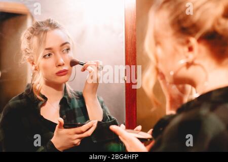 Giovane donna che contorna il suo viso applicando trucco con spazzola trucco guardando se stessa nello specchio a casa prepararsi ad uscire Foto Stock