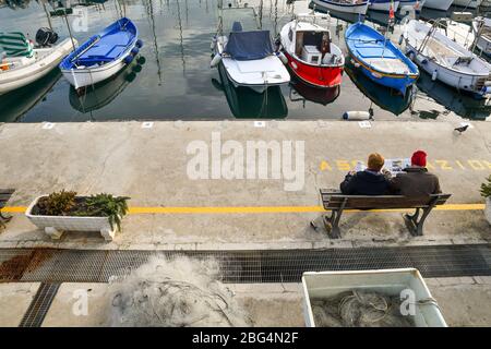 Vista panoramica di una coppia anziana seduta su una panchina e lettura di giornali sulla banchina del porto in una giornata invernale di sole, Sanremo, Liguria Foto Stock