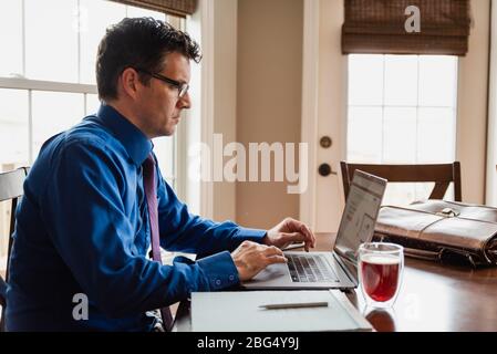Uomo in camicia e cravatta che lavora da casa usando il computer al tavolo da pranzo.