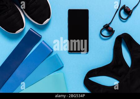 Piatto di reggiseno sportivo, scarpe da running, telefono cellulare e bande di resistenza su sfondo blu. Attrezzature sportive per il fitness a casa. Uno stile di vita sano, Foto Stock