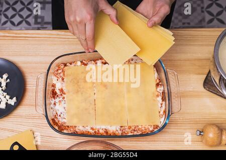 Mani dell'uomo che fanno strati di lasagne in vassoio di vetro, vista dall'alto. Foto Stock