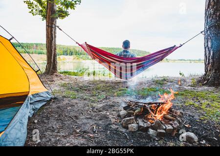 uomo che si posa su amaca sulla spiaggia del lago vicino al fuoco del campo Foto Stock