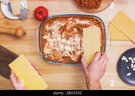 Mani dell'uomo che fanno strati di lasagne in vassoio di vetro, vista dall'alto. Foto Stock