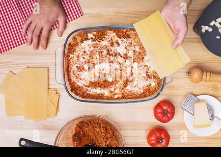 Mani dell'uomo che fanno strati di lasagne in vassoio di vetro, vista dall'alto. Foto Stock