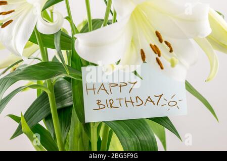 Regalo di fiori freschi per un compleanno con un bouquet di gigli bianchi con una nota scritta a mano che dice buon compleanno, vista primo piano della nota su le verde Foto Stock