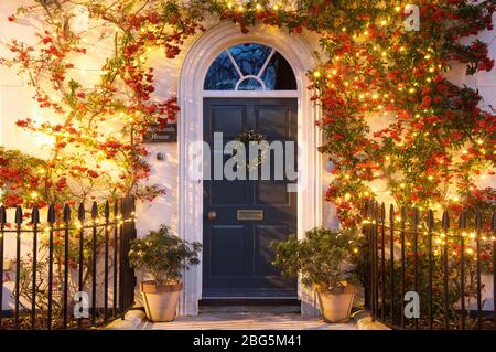 Porta anteriore di una casa incorniciata da una pianta di Pyracantha rampicante decorata con grappoli di bacche rosse e luci di Natale. Poundbury, Dorchester, UK, GB. Foto Stock