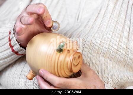 Risparmio, soldi, assicurazione di rendita, pensionamento e concetto della gente - primo piano della mano dell'uomo anziano che mette la moneta nella banca piggy. Foto Stock