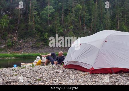 Un giovane uomo bearded beve il tè durante un viaggio in campeggio, in natura, sullo sfondo di una tenda turistica. Foto Stock