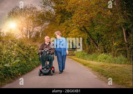 Anziani maturi sposati felicemente coppia fuori nella campagna mentre uno guida uno scooter motorizzato di mobilità. Concetto di assistenza sanitaria Foto Stock