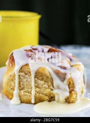 Vista ravvicinata di un rotolo di cannella dal lato. La glassa gocciola lungo il lato con una tazza di caffè sfocata sullo sfondo. Foto Stock