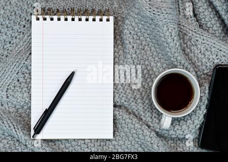 Un'accogliente scrivania a forma di piatto con caffè caldo, carta da appunti e smartphone. Lavoro da casa. Scrittura. Concetto caldo in condizioni climatiche fredde. Foto Stock