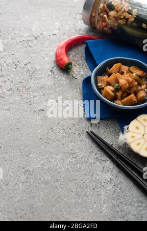 gustosi kimchi in ciotole e vasetto su panno blu vicino a bacchette, peperoncino e aglio a fette su superficie di cemento Foto Stock