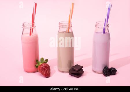 Bottiglie di milkshake con cannucce vicino alla fragola, more e pezzi di cioccolato su rosa Foto Stock