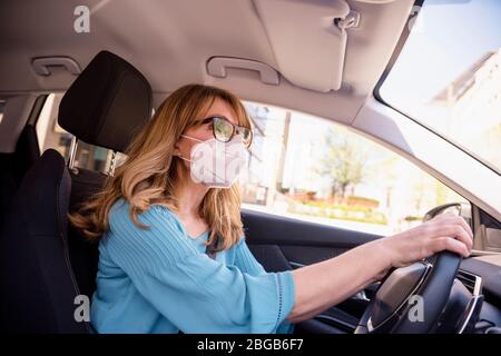 Colpo di donna di mezza età che indossa maschera facciale mentre guida la sua auto durante la pandemia coronavirus. Foto Stock