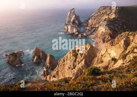 Tramonto sulla spiaggia di Praia da Ursa. Crinale rocciosa con catasta di mare nell'oceano atlantico illuminata da luce calda serale. Vacanze nella regione di Sintra, Portug Foto Stock