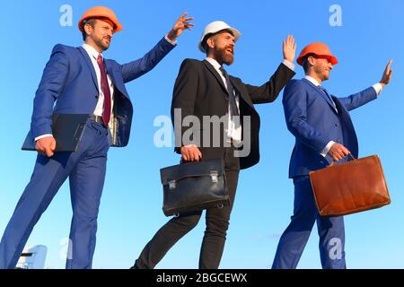 Costruttori a piedi e wave a qualcuno sul cielo blu sullo sfondo. Coworking e costruzione concetto di business. Gli architetti con volti felici in tute e caschi. Gli ingegneri della cartella in attesa, nero e marrone casi Foto Stock